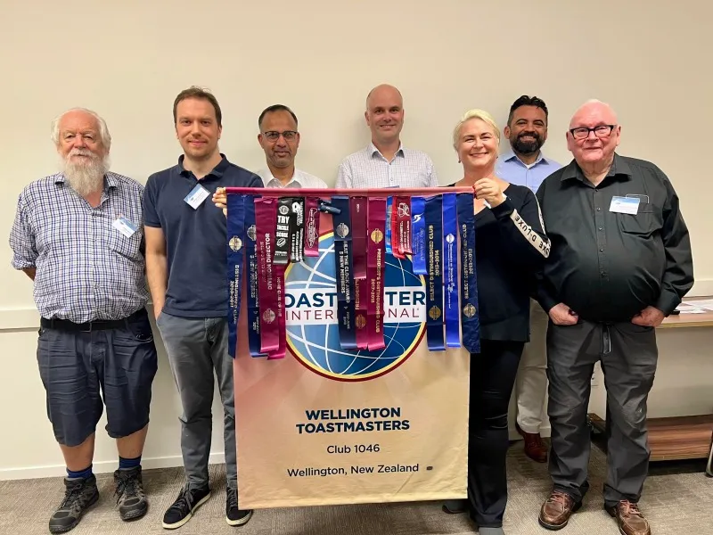 Seven Wellington Toastmasters standing behind the Club 1046 banner